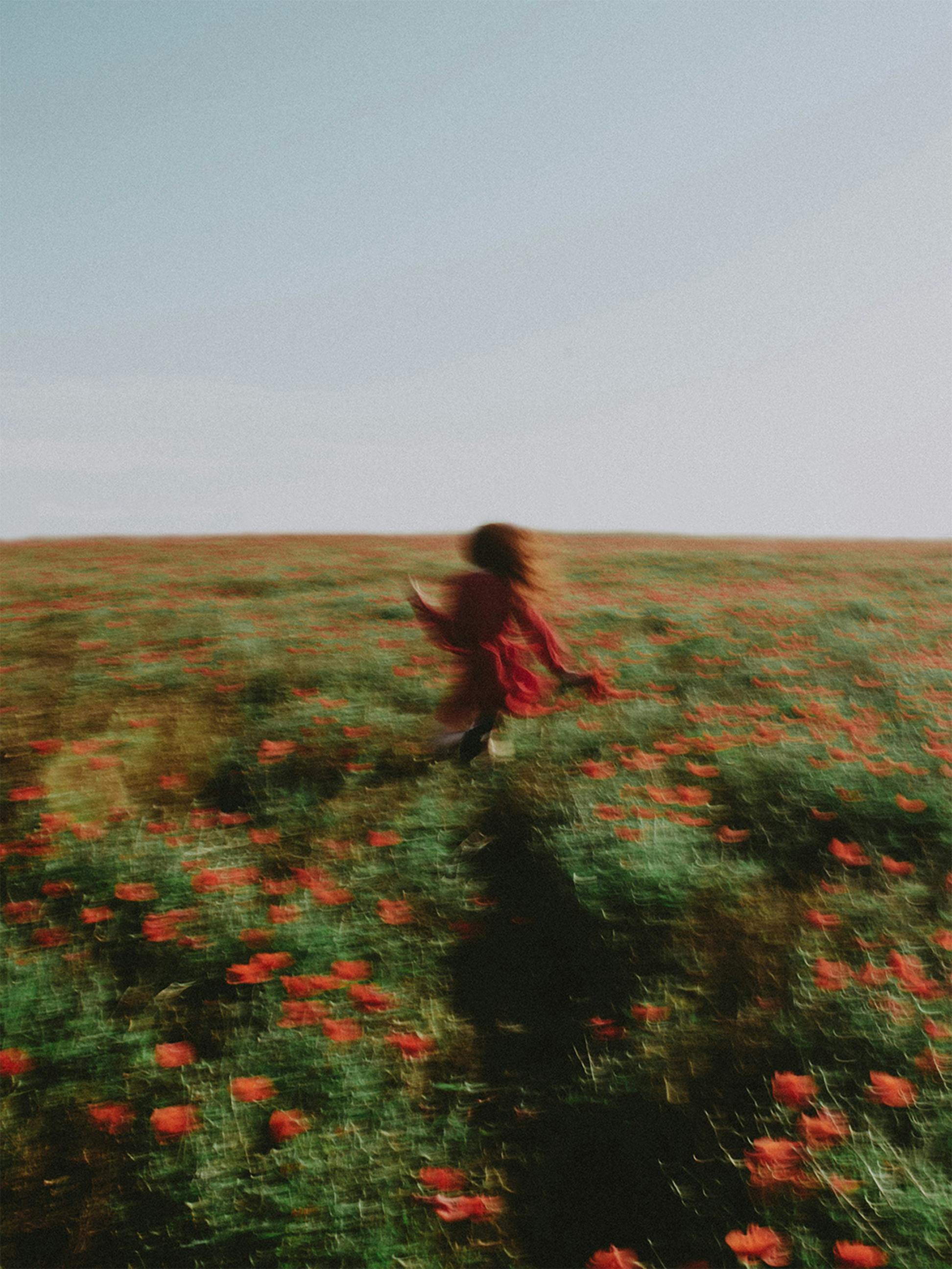 a blurry photo of a person in a red dress runs through a green field with matching red flowers and a very pale blue grey sky.